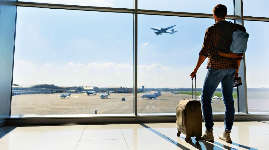 Мужчина-турист с чемоданом и рюкзаком смотрит в окно терминала аэропорта